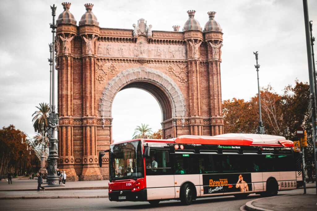 Red city bus passing by the historic Arc de Triomf in Barcelona with autumn leaves.